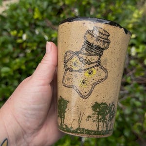 May include: A tan ceramic mug with a black rim and a detailed illustration of a potion bottle. The bottle design includes a cork, rope, and yellow accents. The base of the mug features a green forest scene.