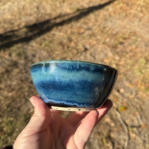 May include: A small, blue ceramic bowl with a white rim. The bowl has a blue and green glaze with a subtle ombre effect.