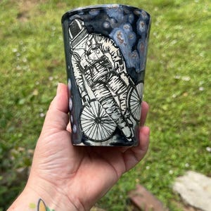 May include: A ceramic tumbler featuring a black and white illustration of an astronaut riding a bicycle. The background of the cup has a dark blue and black mottled glaze with lighter blue circular accents, creating a space-like appearance.