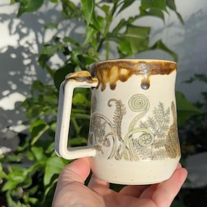 May include: A cream-colored ceramic mug featuring a dark brown drip glaze around the rim and a square-shaped handle. The body of the mug is decorated with detailed botanical illustrations of ferns and fiddleheads in shades of green and brown.
