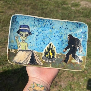 May include: A rectangular ceramic tray featuring a hand-painted scene of a green alien wearing a blue bucket hat behind a tent, a yellow and black campfire, and a large black Bigfoot figure standing on a blue speckled background.