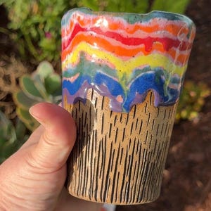 May include: A ceramic tumbler featuring a textured, tan-colored base with vertical carved lines and a vibrant, multi-colored drip glaze at the top. The glaze displays horizontal bands of red, orange, yellow, green, blue, and purple colors.