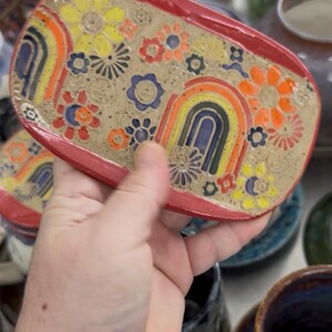 May include: A rectangular ceramic dish with a red rim. The dish is decorated with colorful flowers and rainbows in yellow, orange, blue, and green. The background is a speckled tan color. A hand is holding the dish.