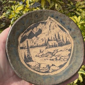 May include: A small, round ceramic dish with a hand-drawn mountain and cabin scene. The rim is a dark blue-green, and the interior features a tan landscape with black mountain and tree details. The dish is held in a hand.