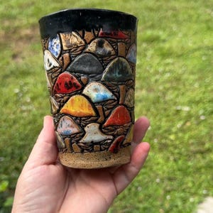 May include: A ceramic tumbler featuring a textured, relief-style surface covered in a dense pattern of mushrooms. The mushrooms are glazed in various colors including red, yellow, blue, and grey, set against a speckled tan clay base with a black rim.