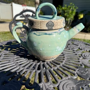 May include: A teal blue ceramic teapot with a lid featuring a seashell design. The teapot has a round body and a spout on the side. It is sitting on a black metal table.