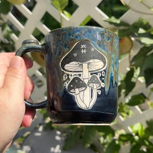 May include: A ceramic mug with a dark blue and brown drip glaze finish. The front features a white graphic illustration of three mushrooms with a Pisces zodiac symbol on the largest cap, surrounded by small star shapes and a smooth handle.