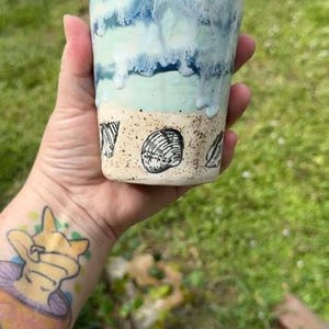 May include: A ceramic tumbler featuring a sandy-colored base decorated with black line drawings of seashells. The upper portion of the vessel displays a layered glaze effect in shades of light blue and deep navy, mimicking ocean waves.