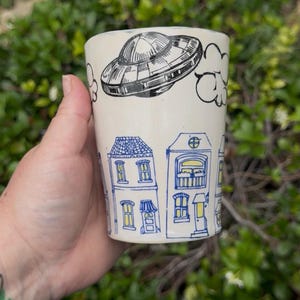 May include: Cream-colored ceramic mug with a hand-drawn design. The mug features a black and white UFO above a row of blue and yellow houses. The design wraps around the mug, creating a whimsical and artistic aesthetic. The mug is held in a hand.