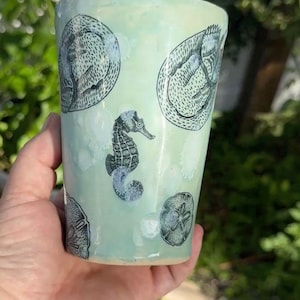 May include: A light mint green ceramic tumbler featuring black ink illustrations of a seahorse, sand dollars, and circular patterns. The cup has a smooth, glossy finish and a tapered cylindrical shape, showcasing detailed marine life imagery.