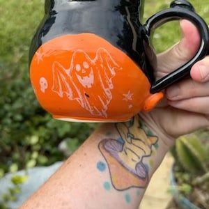 May include: A round ceramic mug with a glossy black upper section and a bright orange lower section. The orange portion features a white etched design of a smiling ghost, a small skull, and several stars. It has a black handle on the right side.