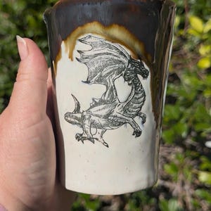 May include: A handmade ceramic mug with a dragon illustration. The mug has a white base with a detailed black dragon drawing. The rim is a dark brown color, with a dripped glaze effect. The mug is held in a hand.
