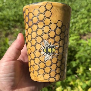 May include: A cylindrical ceramic tumbler with a yellow glaze featuring a black printed honeycomb pattern across the surface. A detailed illustration of a bumblebee with yellow and black stripes and translucent wings is centered on the front.