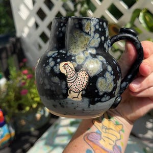 May include: A ceramic pitcher with a rounded body and a curved handle, featuring a black glaze with mottled light green and white crystalline patterns. A small, hand-painted illustration of a chicken with red, black, and white spots is centered.