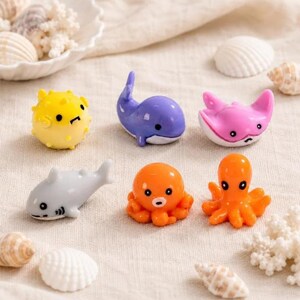 May include: A collection of six small, smooth, glossy resin figurines shaped like sea creatures, including a yellow pufferfish, a purple whale, a pink stingray, a gray shark, an orange octopus, and a second orange octopus figurine.