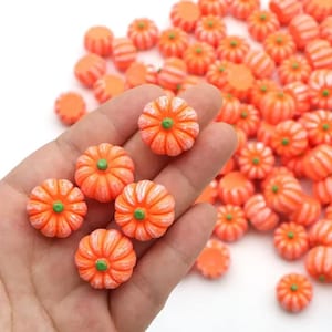 May include: A collection of small, orange pumpkin-shaped beads with white vertical ribbing and a green stem at the centre. These miniature decorative items feature a rounded, segmented design and are scattered across a plain white surface.