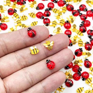May include: A collection of small, flat-backed resin charms shaped like ladybugs and bees. The ladybugs are red with black spots, and the bees are yellow with black stripes and white wings. Several charms are resting on a palm for scale.