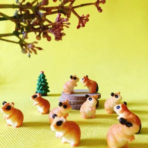 Pode incluir: Uma coleção de pequenas figuras de hamster castanho-claras com olhos e orelhas pretos, dispostas numa superfície amarela. Um par de hamsters está sobre uma pilha de discos circulares perto de uma árvore de Natal verde em miniatura.