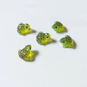 Könnte beinhalten: Fünf kleine, durchscheinende lindgrüne Froschfiguren, die verstreut angeordnet sind. Jeder Frosch hat eine glatte, glänzende Oberfläche mit ausgeprägten dunkelbraunen Flecken auf dem Rücken und Kopf sowie kleinen, soliden schwarzen Augen.