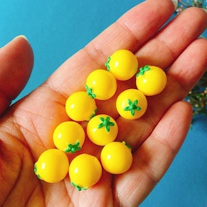 May include: A collection of ten small, round, bright yellow beads shaped like miniature tomatoes. Each bead features a smooth, glossy finish and a small, moulded green star-shaped stem attached to the top, creating a uniform, fruit-inspired design.