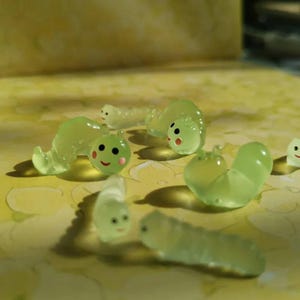 Può includere: Una collezione di piccole statuette di bruchi in resina verde chiaro traslucido. Ogni statuetta presenta un corpo liscio e segmentato, due piccole antenne e un volto dipinto con occhi neri, un sorriso rosso e guance rosa.