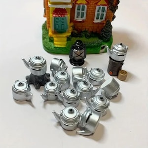 Könnte beinhalten: Eine Sammlung von silberfarbenen Miniatur-Metallkesseln, die vor einem kleinen, detaillierten Modell eines Backsteinhauses angeordnet sind. Das Set enthält eine schwarze Laterne, einen braunen Ofen mit einem Kessel darauf und mehrere Kessel.