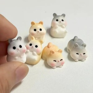 May include: A collection of six miniature hamster figurines in various poses, featuring soft color palettes of white, light gray, and pale orange. Each small animal has rounded features, black eyes, and pink cheeks, crafted with a smooth finish.