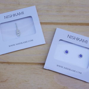 Może przedstawiać: Dwa małe białe pudełka z tekstem "NISHKAMI" i adresem strony internetowej "www.nishkami.com". Pudełka zawierają dwa różne rodzaje biżuterii: srebrny wisiorek w kształcie łzy i parę małych niebieskich kolczyków z kwiatowym wzorem.