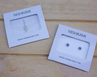 May include: Two small white boxes with the text "NISHKAMI" and the website address "www.nishkami.com". The boxes contain two different types of jewelry: a silver pendant with a teardrop design and a pair of small blue earrings with a flower design.