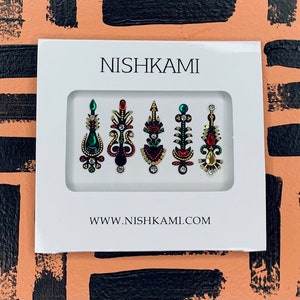 May include: A white rectangular package with the brand name "NISHKAMI" and website address. The package contains six colorful, jewel-toned bindi stickers in various ornate designs. The background is a peach color with black brush strokes.