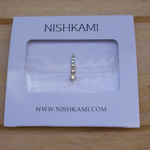 May include: A white card with the brand name "NISHKAMI" and website address. The card features a clear window showcasing a decorative forehead jewel with iridescent gemstones in a vertical design.