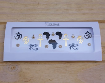 May include: A white card with a clear plastic window showing a set of black and gold temporary tattoos. The tattoos include the Om symbol, an ankh, an eye of Horus, an African continent outline, and a symbol that looks like a stylized letter 'A'. The card also has the text "NISHKAMI" in black.