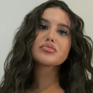 Peut inclure: Gros plan d'une personne aux longs cheveux ondulés foncés et aux lèvres pulpeuses. Elle porte un haut noir avec un décolleté plongeant et un détail de laçage.