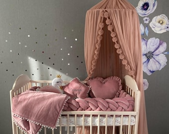 May include: A white wooden crib with a pink and white bedding set. The crib has a pink canopy with pom poms around the edge. The bedding includes a pink blanket, a pink pillow with the word "Princess" embroidered on it, and a pink braided bumper.
