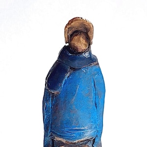 Peut inclure: Petite sculpture artisanale d'une personne vêtue d'un manteau bleu et d'un chapeau. La sculpture a une surface texturée et un aspect rustique. Le chapeau et le visage sont de couleur bronze, contrastant avec le bleu du manteau.