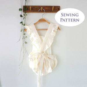 Puede incluir: Un pelele de bebé de lino blanco con tirantes con volantes y un lazo en la cintura. El pelele está colgado de una percha de madera contra una pared blanca. El texto "Sewing Pattern" está escrito en un óvalo azul en el lado derecho de la imagen.