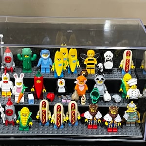 May include: A clear plastic display case with two shelves holds a collection of colorful Lego minifigures. The minifigures are dressed in various costumes, including a hot dog, a banana, a penguin, a cactus, and a king.