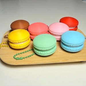 以下が含まれることがあります： 茶色、赤、ピンク、黄色、緑、青の色合いの、カラフルな丸いマカロンのコレクション。各マカロンは木製のトレイにあり、一部にはキーチェーンが付いています。マカロンはデザートをテーマにしたアイテムです。