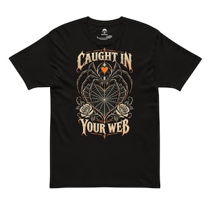 May include: A black short-sleeved t-shirt featuring a graphic of a spider above a heart-shaped web, flanked by two roses. The text "CAUGHT IN YOUR WEB" is printed in a stylised font above and below the central illustration in cream and orange.