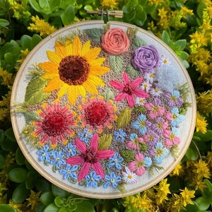 Pode incluir: Um aro de bordado colorido que apresenta uma variedade de flores, incluindo um grande girassol, flores rosa e vermelhas e miolos-de-ouro azuis.