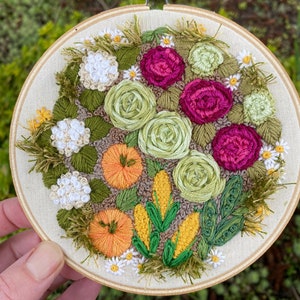 May include: Embroidered hoop art featuring a floral and vegetable garden scene. The design includes pink roses, green cabbage roses, white hydrangeas, orange pumpkins, yellow corn, and green leaves.