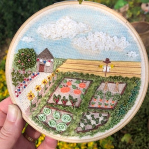 May include: Embroidered hoop art depicting a whimsical garden scene with a small house, fluffy clouds, and a variety of flowers and vegetables. The hoop is made of wood and has a natural finish.