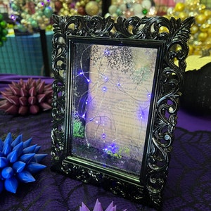 May include: A black ornate picture frame with a glass front. The frame contains a piece of paper with text and a string of blue fairy lights. The frame is sitting on a purple table cloth.