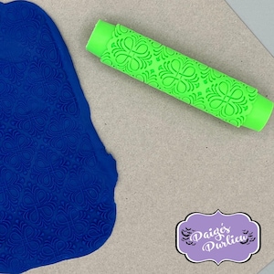 May include: A bright green polymer clay roller with a floral pattern, next to a piece of blue clay with the same embossed design. The roller is cylindrical with a textured surface. The image also includes a logo with the text "Paige's Purlieu".