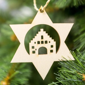 Puede incluir: Un adorno de Navidad de madera en forma de estrella con un diseño recortado de un edificio en su interior. El adorno está colgado de un árbol de Navidad verde.