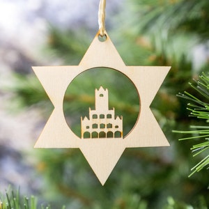 Puede incluir: Un adorno de Navidad de madera en forma de estrella con un recorte de un edificio en el centro. El adorno está colgado de un árbol de Navidad.
