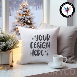 Pode incluir: Uma almofada branca com o texto "YOUR DESIGN HERE" em letras pretas e algumas pequenas estrelas. A almofada está sobre uma superfície branca com uma almofada cinza atrás. Uma xícara de café branca e um pires estão sobre a superfície à direita da almofada.