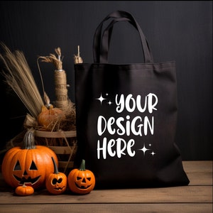 May include: Black tote bag with white text that says "YOUR DESIGN HERE" with stars on either side of the text. The bag is sitting on a wooden surface with three small orange pumpkins in front of it.
