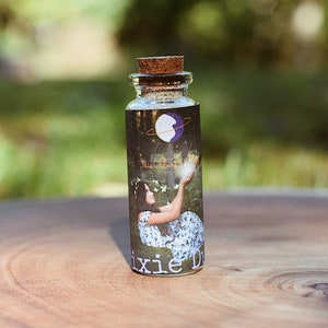 May include: A small glass bottle with a cork stopper and a label featuring a woman holding a glowing orb. The label also includes the text "Dixie Dust".