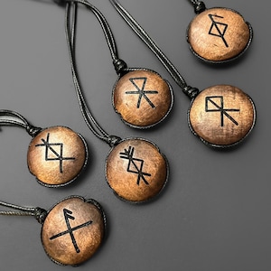 Puede incluir: Una colección de collares con colgantes de madera y cordones negros. Cada colgante es un disco circular de color bronce con un símbolo rúnico negro único. Los colgantes están dispuestos sobre una superficie gris, mostrando sus intrincados diseños.